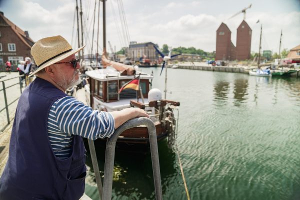 Axel Prahl steht am Hafen und blickt auf das Wasser. Er trägt Sonnenbrille und Sonnenhut.