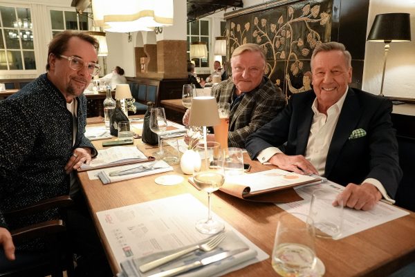 Jan Josef Liefers, Axel Prahl und Roland Kaiser sitzen am Tisch in einem Restaurant und lächeln in die Kamera.