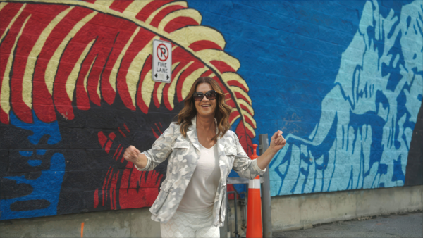 Katarina Witt steht in Calgary vor einer bunt angemalten Mauer und tanzt zur Straßenmusik.